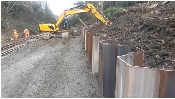 Road rail vehicle removes material from base of slope and replaces with granular material behind sheet piling