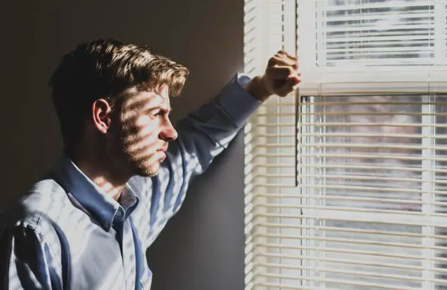 depressed man looking out of window through venetian blinds by Ethan Sykes