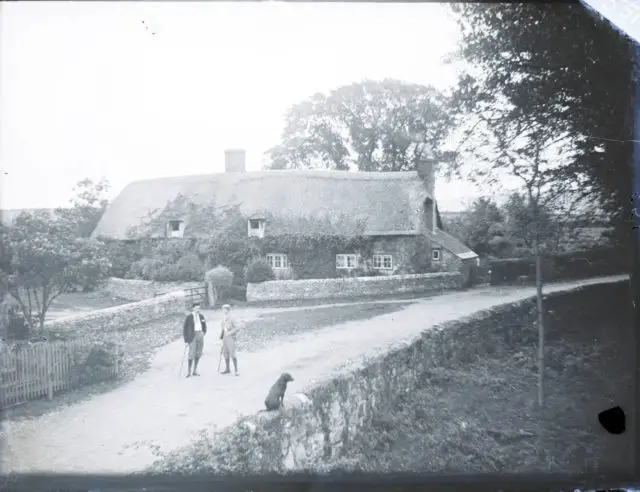 Men chatting outside thatched cottage with dog on the wall