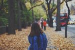 young woman with backpack on walking down tree lined street with red busy in background and autumn leaves on the pavement by Jake Ingle