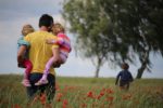 man holding two young girls whilst boy running in poppy field by Juliane Liebermann