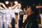 woman drinking from christmas themed cup at christmas fair with fairy lights in background