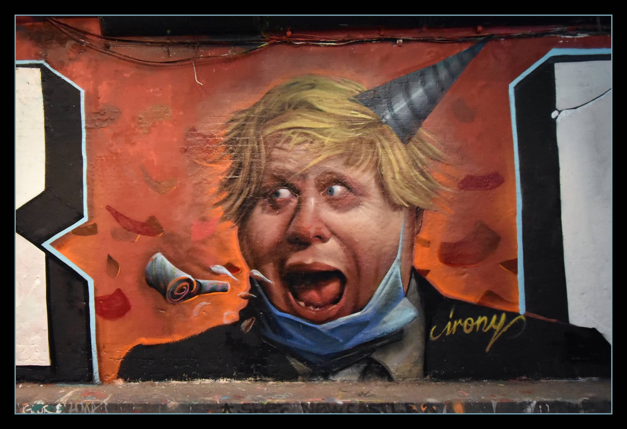 Mural of Boris Johnson with a party hat on, mask around his chin and blowing a party whistle