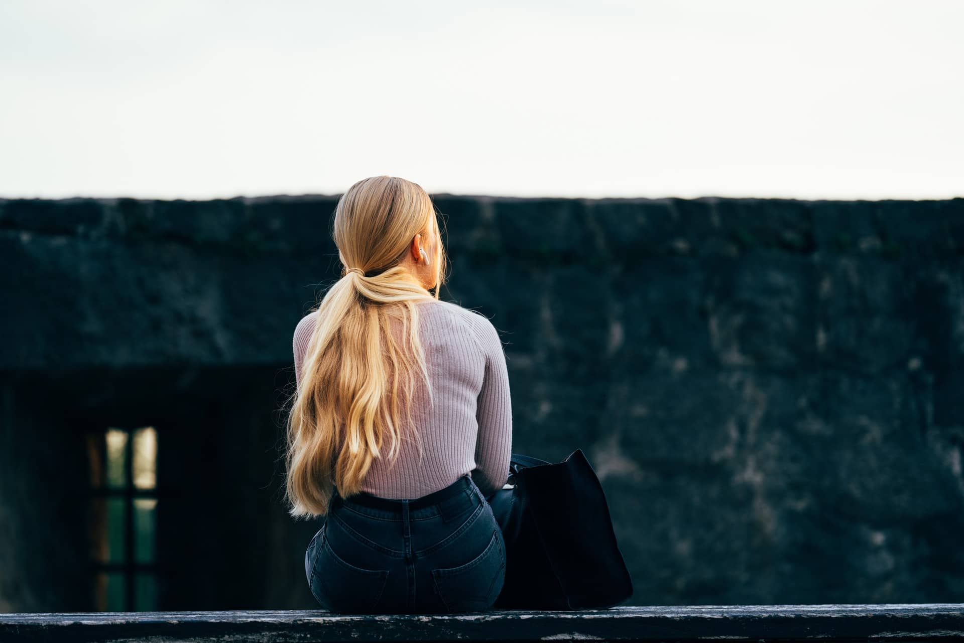 Young female with long blond hair sitting on wall with back to the camera