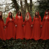 The Mediaeval Baebes dressed in red dresses and cloaks standing hand in hand in woodland