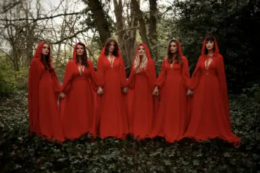 The Mediaeval Baebes dressed in red dresses and cloaks standing hand in hand in woodland