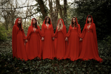 The Mediaeval Baebes dressed in red dresses and cloaks standing hand in hand in woodland