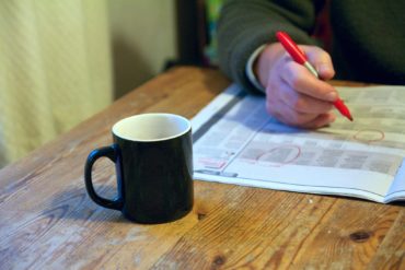 Person marking job ads in a paper, whilst sat at table with mug of tea