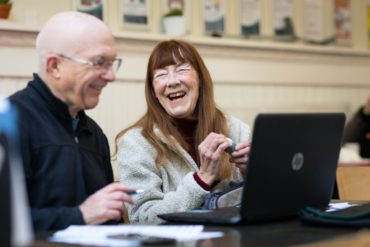 two older people sitting in front of laptops