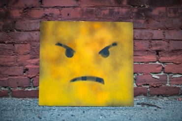 Angry face on yellow board leaning up against a brick wall