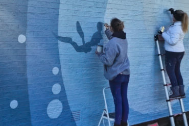 Artist painting the side of Waterisde Pool building for the Ryde Arts Trail