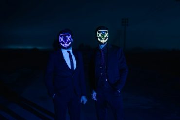 Two men standing in dark wearing neon masks