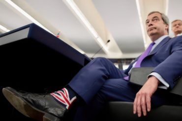 Nigel Farage sitting in a lounge chair, wearing union flag socks by J.M Executive