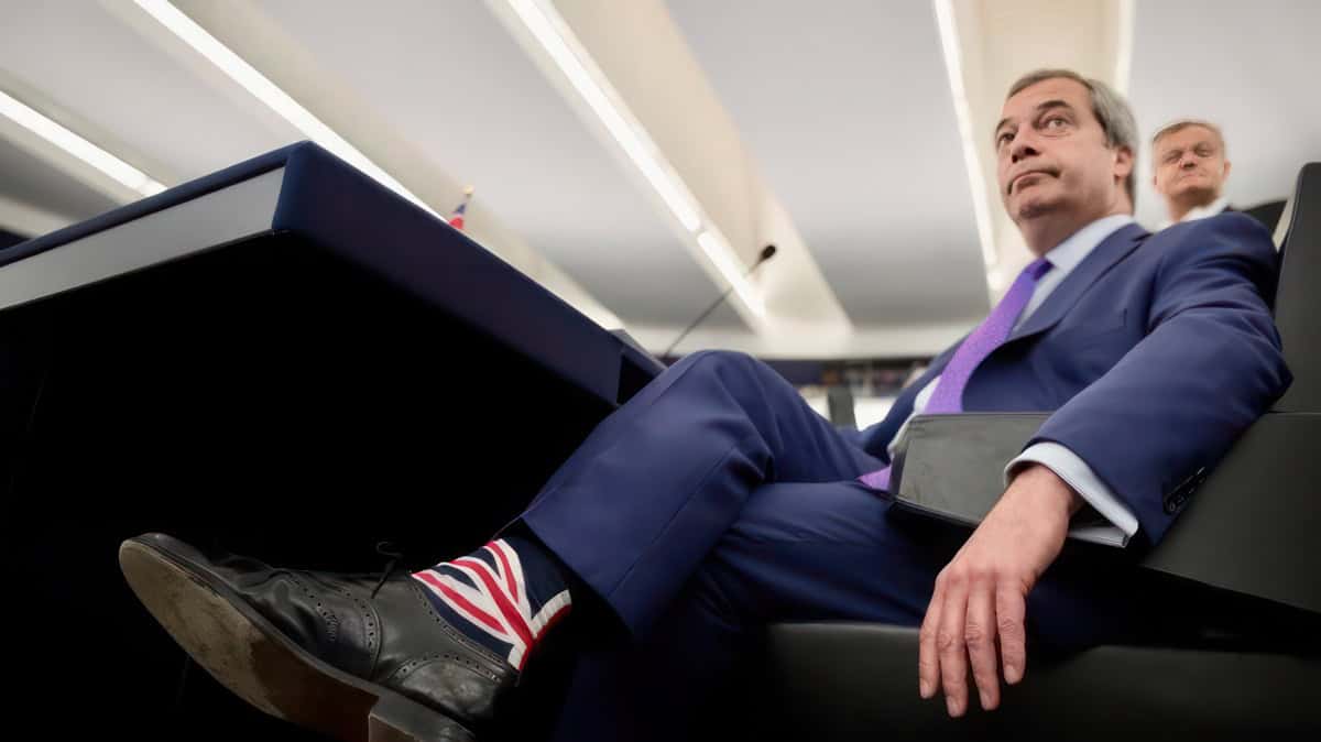 Nigel Farage sitting in a lounge chair, wearing union flag socks by J.M Executive