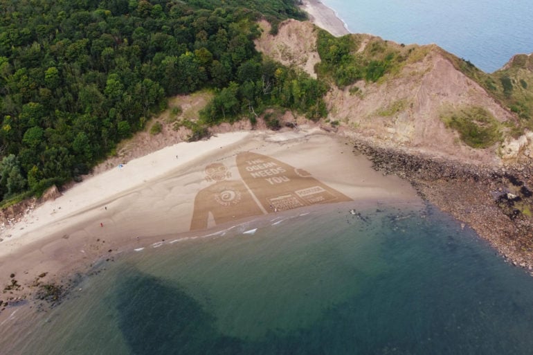 Aerial view of sand art - showing person with gas mask holding a surfboard with the word 'the ocean needs you'