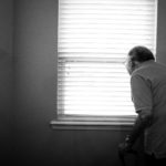 Black and white photo of an older person looking through venetian blinds by alex boyd