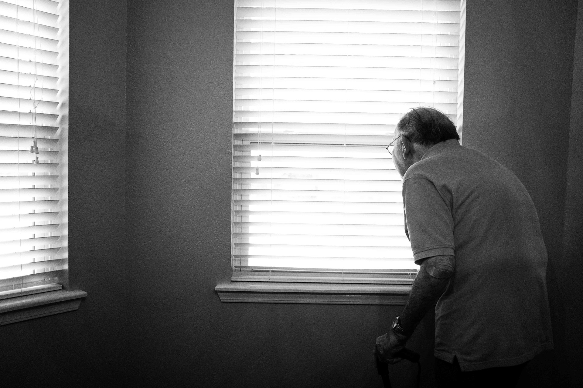 Black and white photo of an older person looking through venetian blinds by alex boyd