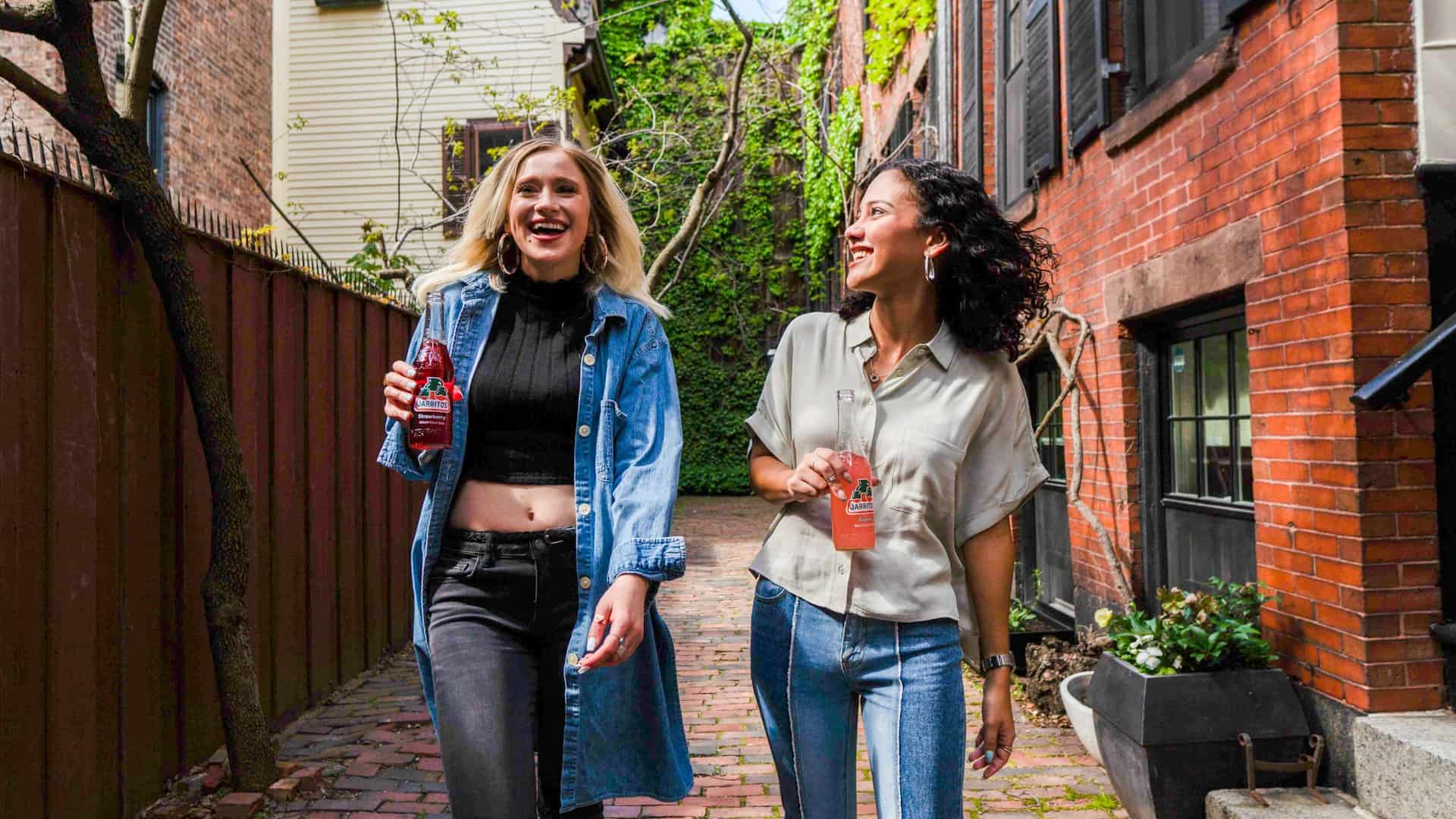 Two young women walking down a street holding glass bottles of pop