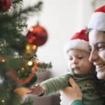 woman and baby looking at a christmas tree, both wearing santa hats