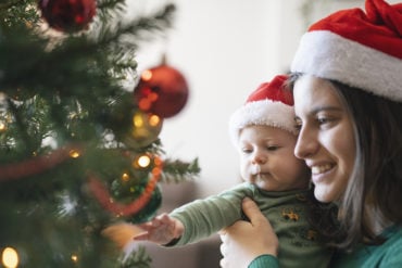 woman and baby looking at a christmas tree, both wearing santa hats