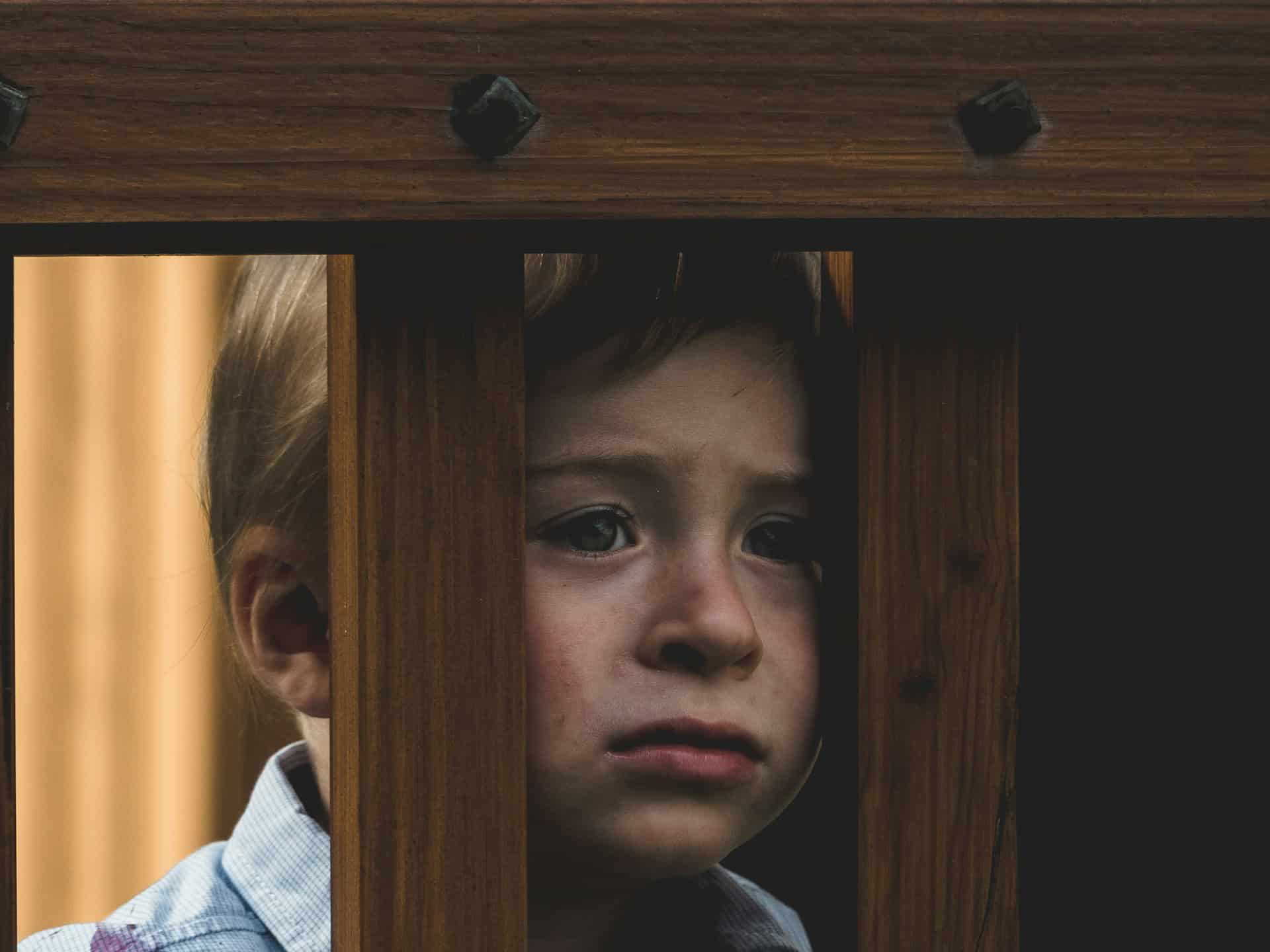 unhappy sad child looking through wood slats of play equipment