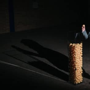 Person standing on dark stage putting plastic balls into a large glass vessel