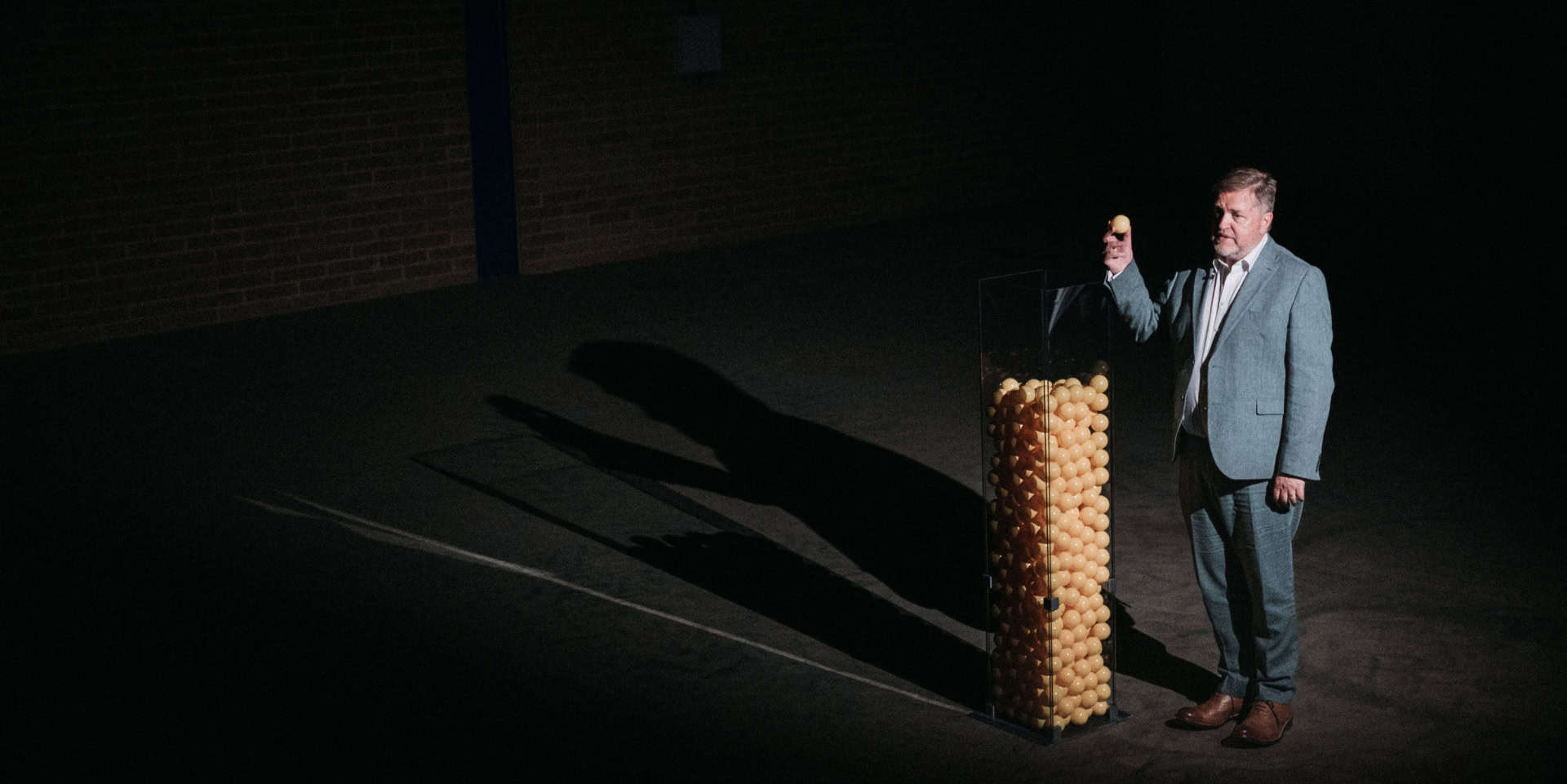 Person standing on dark stage putting plastic balls into a large glass vessel