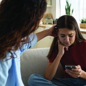 Supportive mother comforting sad teenage girl rejected by friends, helping depressed teen daughter to deal with breakup. Upset depressed adolescent child looking at phone at home, waiting for call