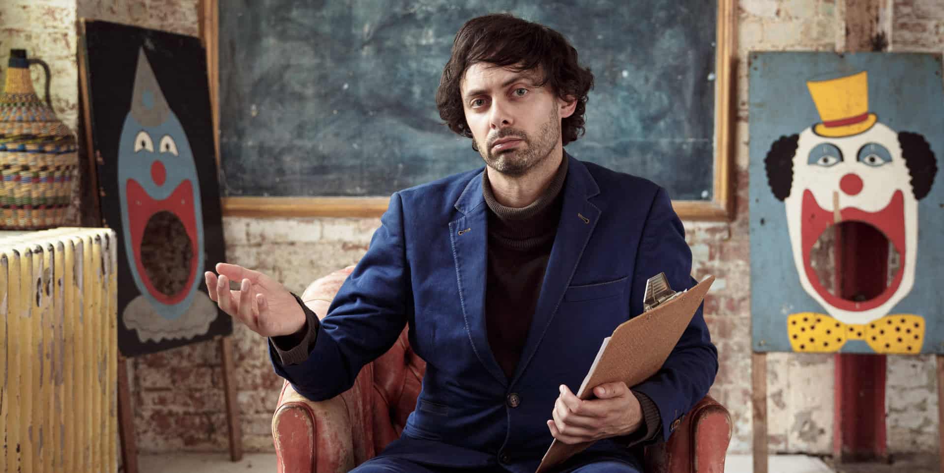 Marcel Lucont sitting in a chair with clown paintings behind him