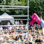 Aerial artist performing on a scaffold tower in front of audience sat on grass in park
