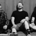 The three band members sitting against a metal container