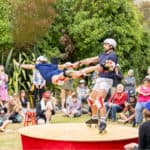 Two circus performers in the Botanic Garden on roller skates with the man swinging the woman in the air, with lots of onlookers