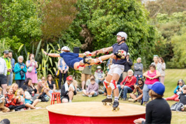 Two circus performers in the Botanic Garden on roller skates with the man swinging the woman in the air, with lots of onlookers