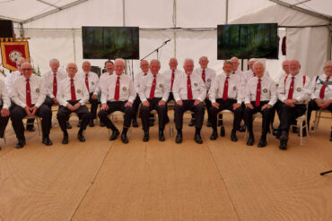The choir, all dressed in black trousers, white shirt and red ties, seated in marquee and about to perform