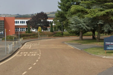 Photo of the school from the road, with lots of trees in the foreground and Downs in the background