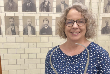 Dr Angela Buckley posing for the camera with Victorian mugshots on the wall behind her