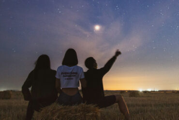 three people sat on hale bales in a field looking at the stars