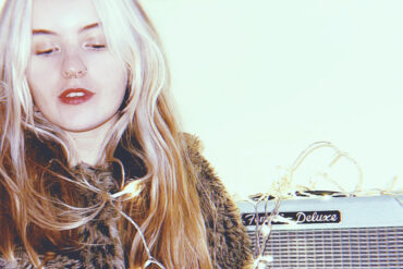 woman with leopard skin fluffy jacket on sitting next to guitar AMP