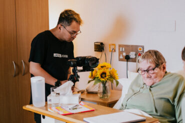 Older woman in hospice bed with grandchild next to her bed and man filming them together