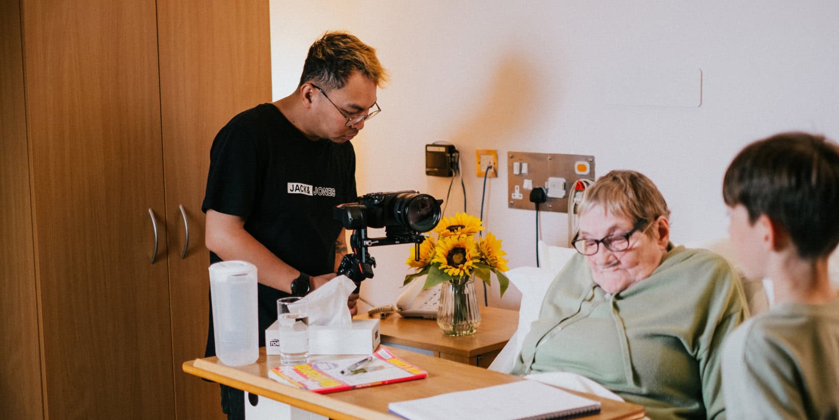 Older woman in hospice bed with grandchild next to her bed and man filming them together