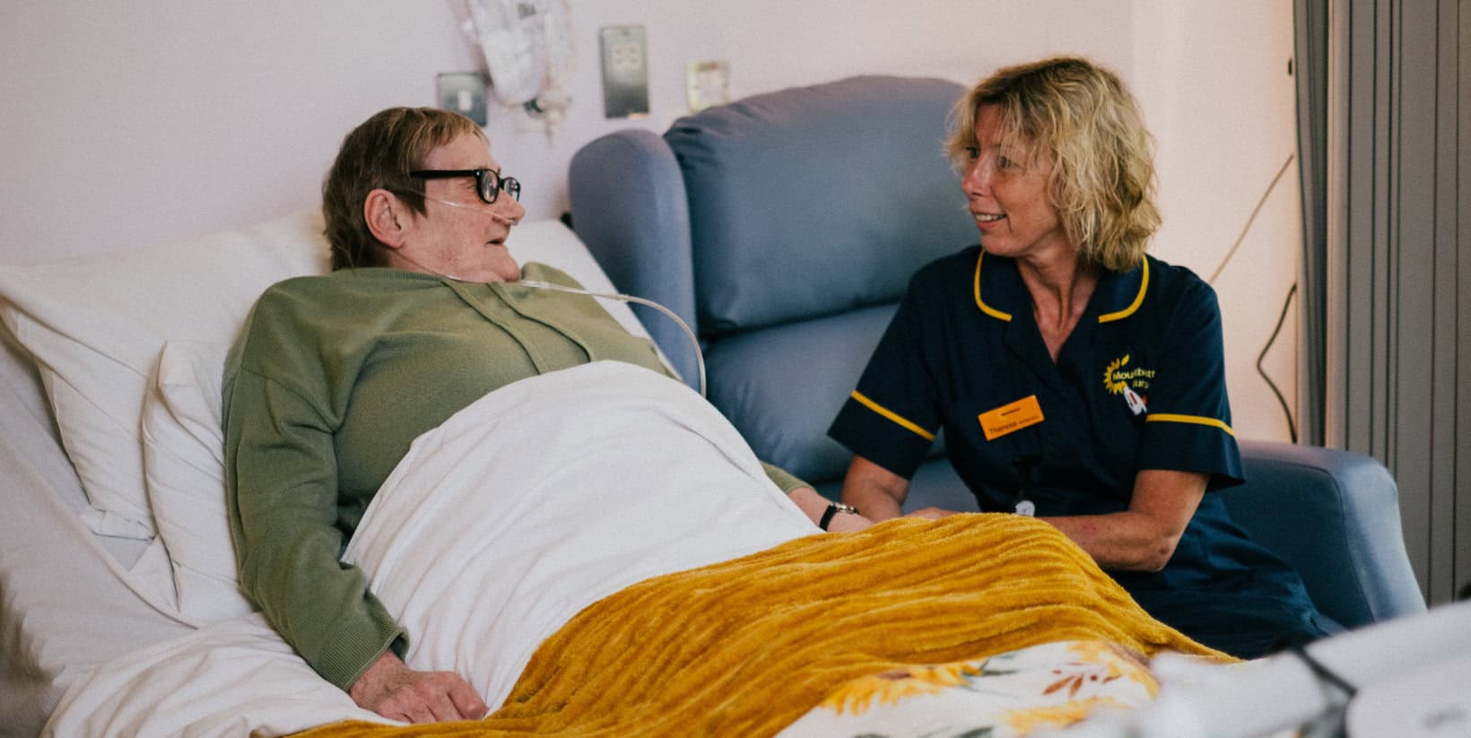 Older woman in hospice bed with Mountbatten nurse sat next to her listening