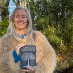 Jane Giles standing outside, wearing a fluffy beige jacket and holding her book