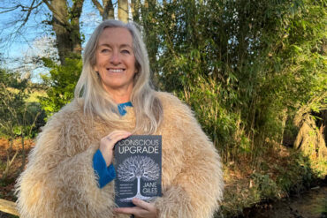 Jane Giles standing outside, wearing a fluffy beige jacket and holding her book