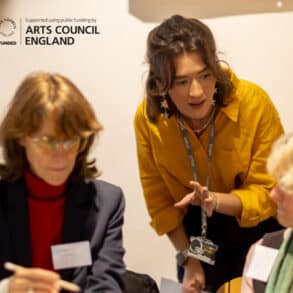 Women sat in on chairs in a workshop setting with a younger woman leaning in to offer help