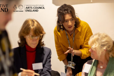 Women sat in on chairs in a workshop setting with a younger woman leaning in to offer help