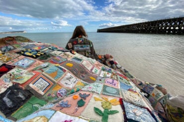 Person wearing the coat of hopes on a beach, with people holding up the coat tails