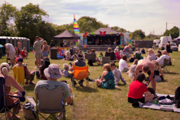 People sitting on the gras or on fold up chairs watching performers on the stage at Three Rivers Music Festival