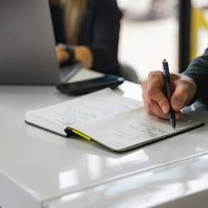Man writing in a notebook whilst sat next to someone using a laptop