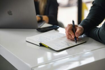 Man writing in a notebook whilst sat next to someone using a laptop