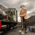 Paul Merton standing next to a steam train and coal pile with shovel in hand